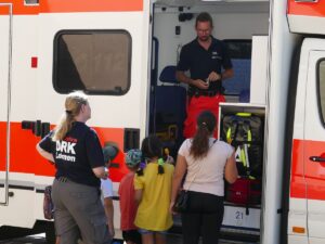 Kinder mit Betreuer*innen vor einem geöffneten Rettungswagen, ein Sanitäter erklärt die Ausstattung.