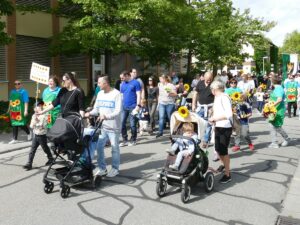 Familien mit Kinderwagen und Sonnenblumen, begleitet von Kindergarten-Team beim Umzug.