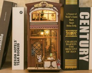 Ein beleuchteter Book Nook in Form eines kleinen Cafés steht zwischen Büchern im Regal; eine detailreiche Miniaturwelt mit Tisch, Stuhl und Dekoration, die zeigt, wie solche Miniaturwelten Kreativität und Konzentration beim Lernen fördern können.