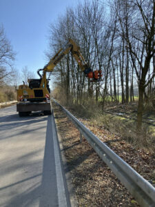 Gelber Gehölzbagger schneidet Gehölze entlang einer Straße zurück.