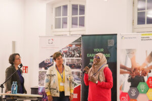 Frau mit Kopftuch hält Mikrofon und spricht vor Publikum.