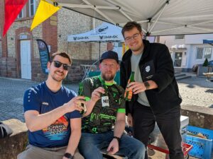 Drei Männer stoßen unter einem Pavillon beim Wochenmarkt in St. Ilgen mit Getränkeflaschen an.