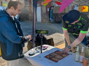 Zwei Männer stehen an einem Tisch mit digitaler Stoppuhr und Getränkeflaschen beim Wochenmarkt in St. Ilgen.