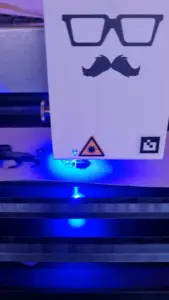 Ein Laserschneider bearbeitet unter blauem Licht ein kleines Materialstück mit ausgeschnittenen Formen.