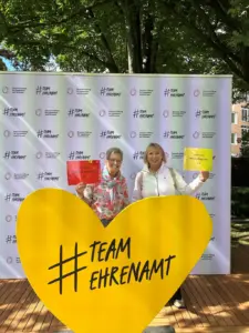 Zwei Frauen halten Vereinskarten der Leimener Mittwochsgruppe vor einem Banner der Deutschen Stiftung für Engagement und Ehrenamt; im Vordergrund ein gelbes Herz mit der Aufschrift Team Ehrenamt.